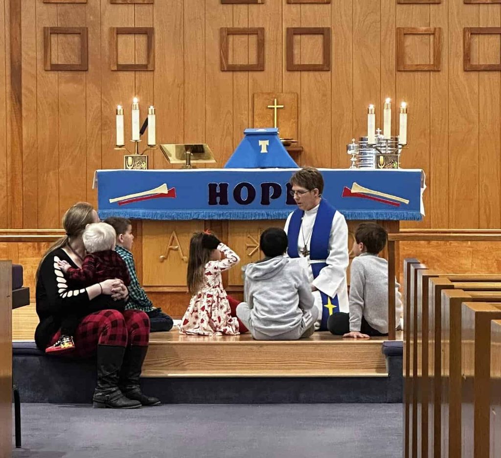 Pastor Chris sits with a group of 5 children and one adult female at the altar during the children's message at Faith Lutheran Church, Bristol CT.