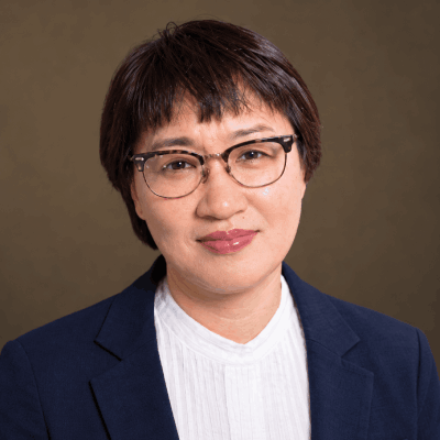 Ms. Lee, the music director, has brown hair and glasses. She is wearing a white shirt with a black jacket.
