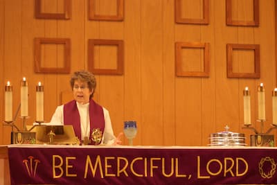 Pastor Chris is at the altar with the wine in the bread and wine. Three candles are lit on each side of the altar. There is a red banner in front of the altar that reads Be Merciful, Lord in lighter colored letters.
