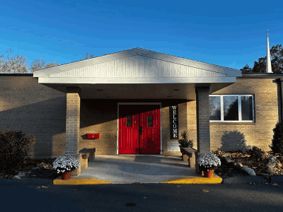 Exterior of Faith Lutheran Church next to parking lot, featuring red entrance doors, a covered entrance, brick walls, and landscaped planters under a clear blue sky.