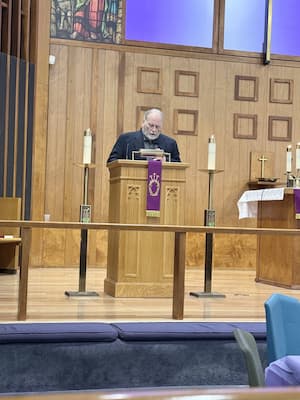 Male person reading from the scripture at the altar. Two high candles are on either side of the lector's table, with a purple banner hanging in front of it.