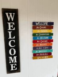 A large black wooden sign on the left that says WELCOME. On the left is a series of smaller signs below the word WELCOME: All Colors, All Genders, All Orientations, All Religions, All Cultures, All Abilities, All Ages - All Sizes, All Individuals, All Means All, Love Lives Here.